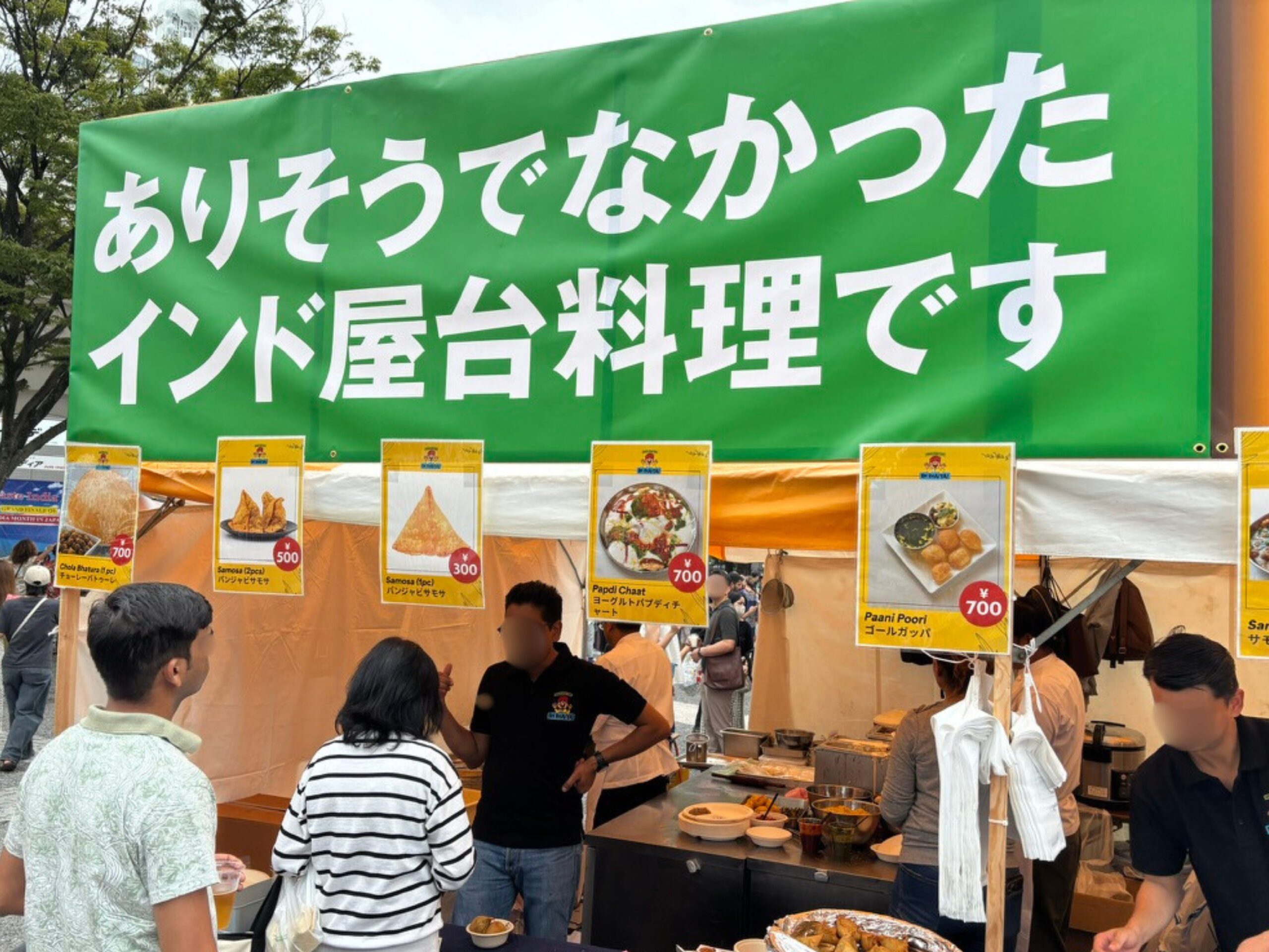「ありそうでなかったインド屋台料理です」と大きく書かれた緑色の看板が目を引くブース。サモサやゴールガッパ(Paani Poori)など本格的なストリートフードを提供し、来場者と談笑するスタッフの姿が写る。東京の国際フェスで撮影。