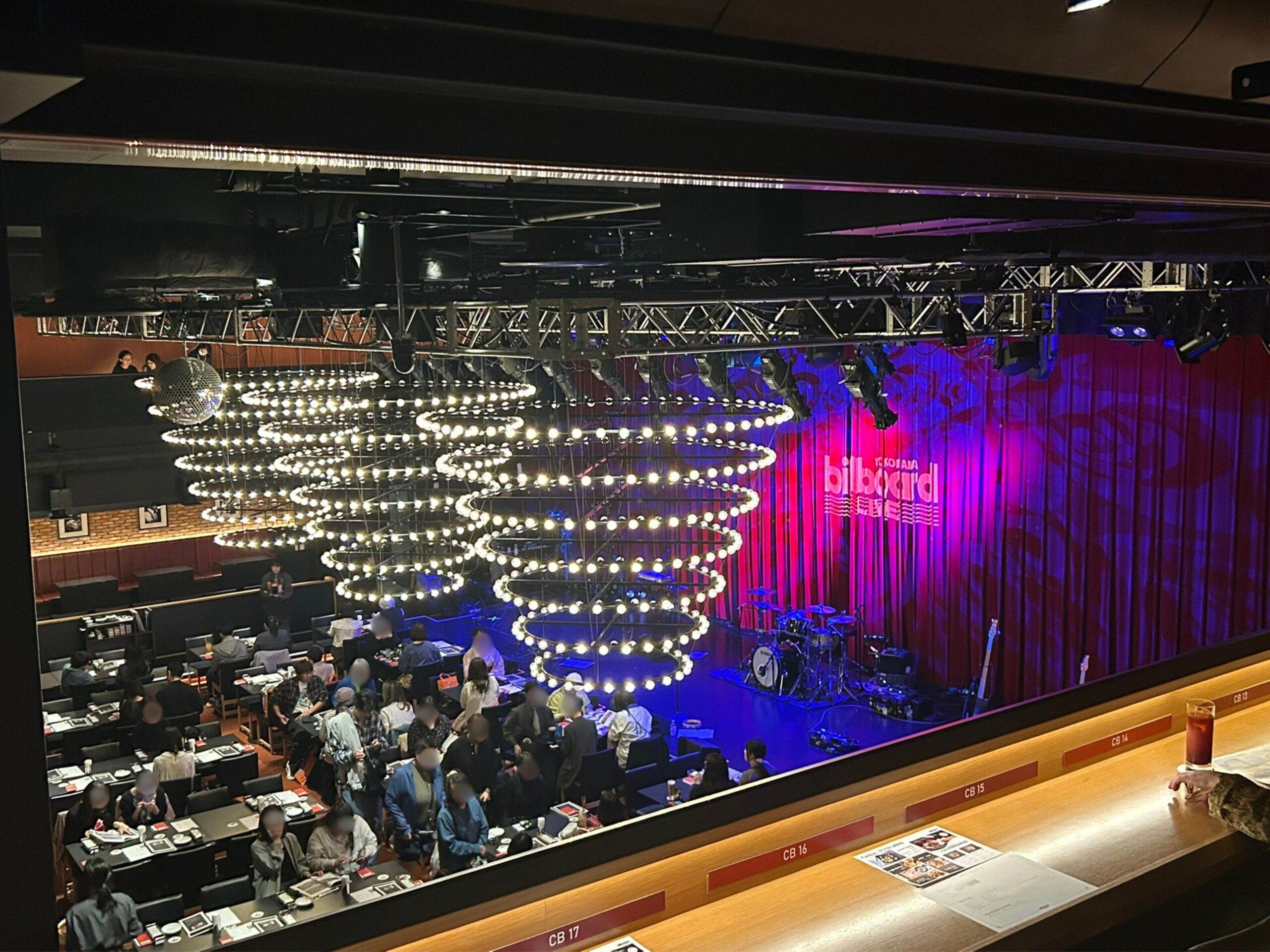 「横浜のBillboard Liveの夜のエントランス。青いネオンライトが輝くガラス張りの入口には、訪れる人々の姿も映る。」開演前室内
