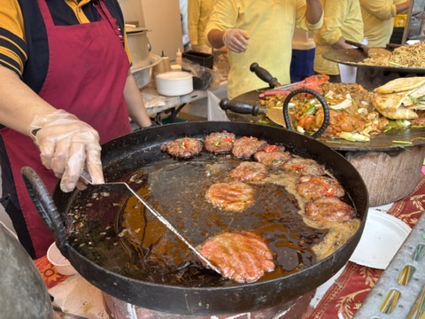 屋台で大きな鉄鍋を使い、肉肉しいチャップリケバブ。(イスラムハンバーグ)をスタッフが調理している様子。赤エプロンの人物がトングで一枚を返している。