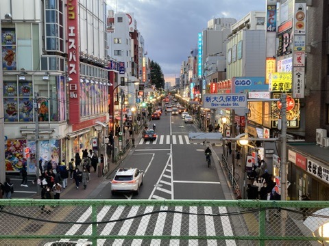 夕暮れ時の新大久保駅前の風景。左右にネオン看板が並ぶ繁華街を車と人が行き交い、活気ある雰囲気が広がる。高架からの俯瞰アングルで撮影。