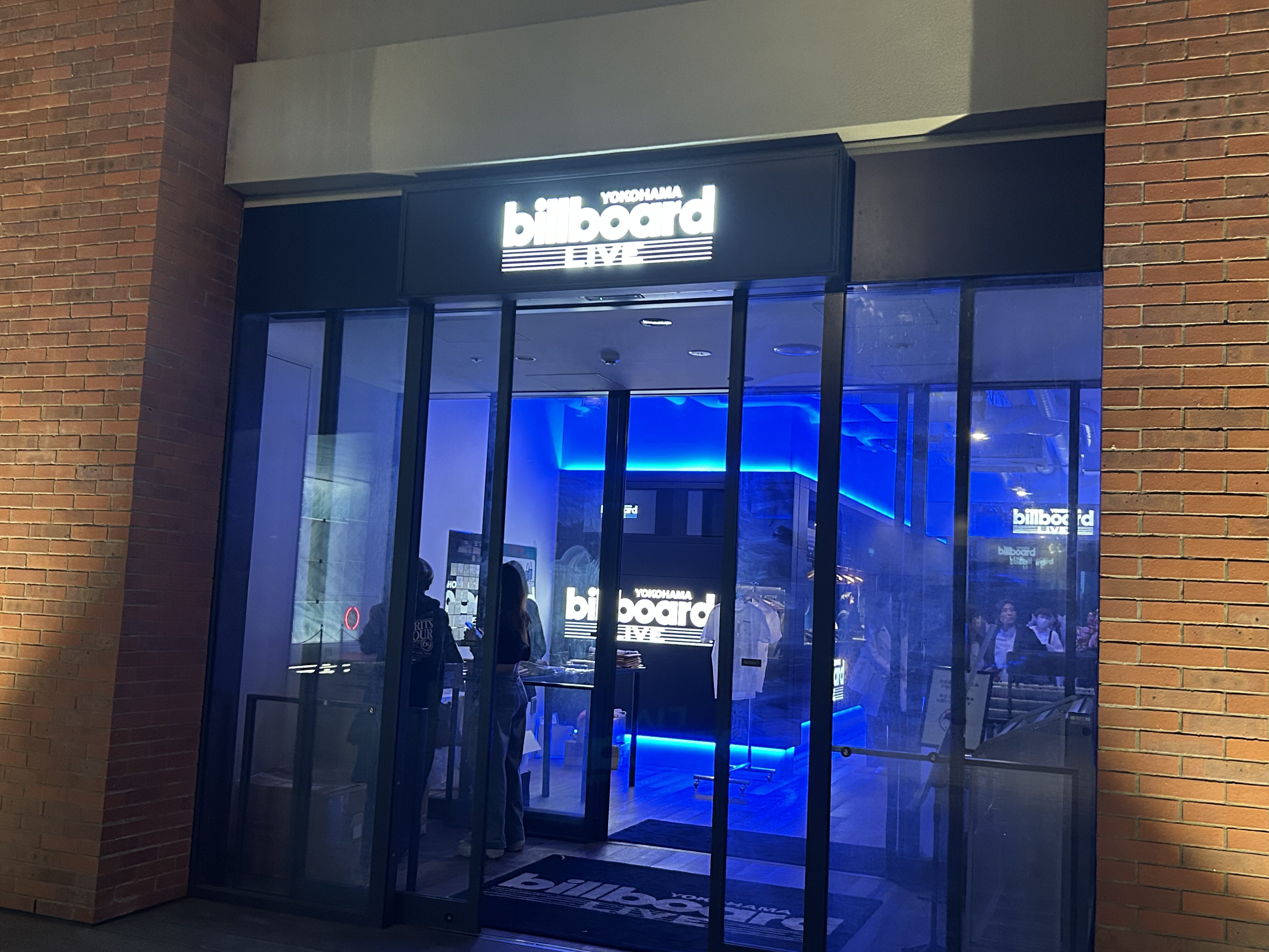 「横浜のBillboard Liveの夜のエントランス。青いネオンライトが輝くガラス張りの入口には、訪れる人々の姿も映る。」