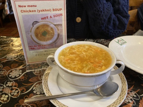 「白い取っ手付きスープボウルに注がれた黄金色のチキンヤクニ。湯気が立ち上り、背景にはメニューと空の白い皿が置かれている」