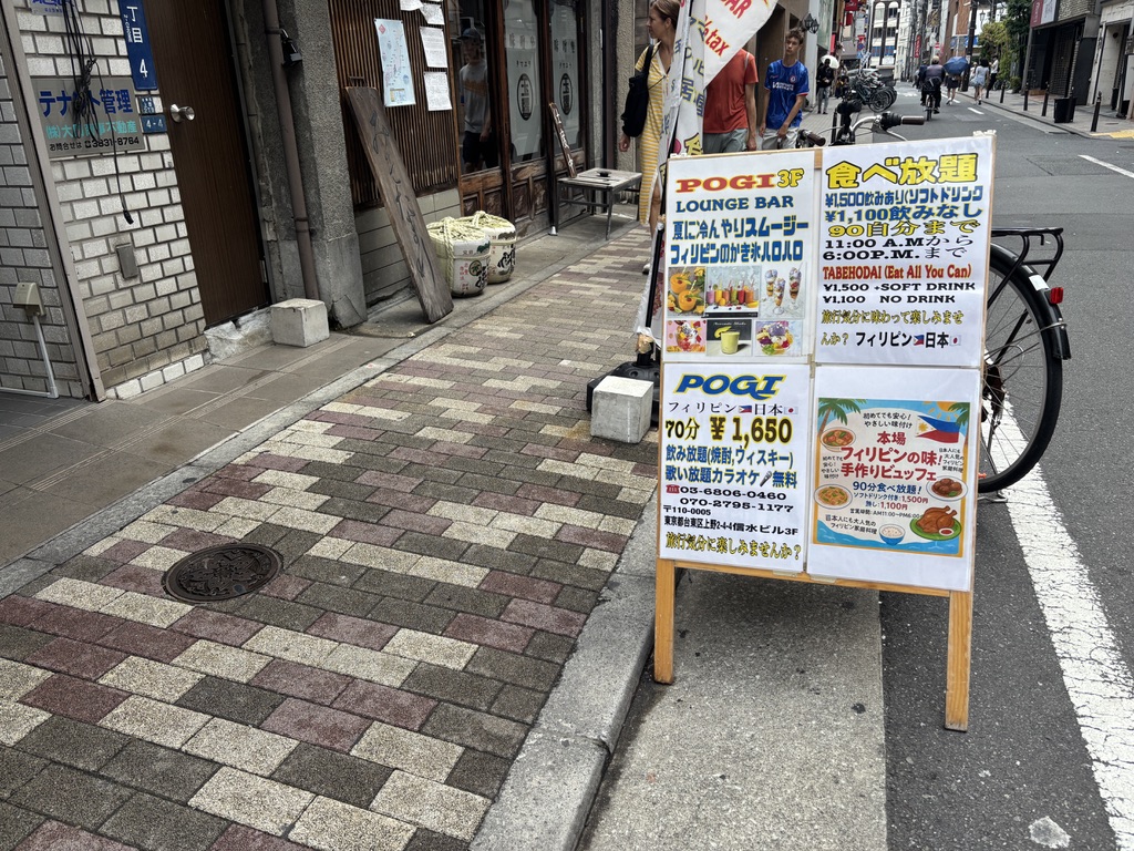  雑居ビル前に設置された、フィリピン料理食べ放題「POGI」の立て看板。ビュッフェの内容や価格が記載され、通行人にも目立つデザイン。
