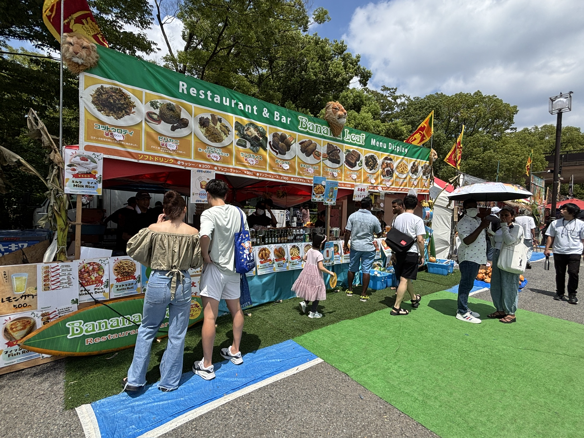屋外イベント会場のスリランカ料理屋台「Banana Leaf」。色鮮やかなメニュー写真が並び、客が注文を待つ様子が見られる。