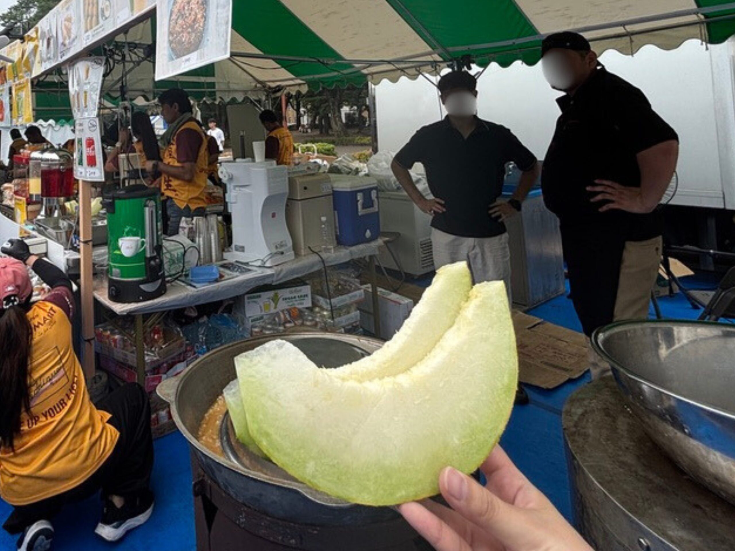 パキスタンフェスティバルの屋台と、ウズベキスタン産メロンを持つ手  
Food stall at Pakistan Festival with a hand holding Uzbek melon slice