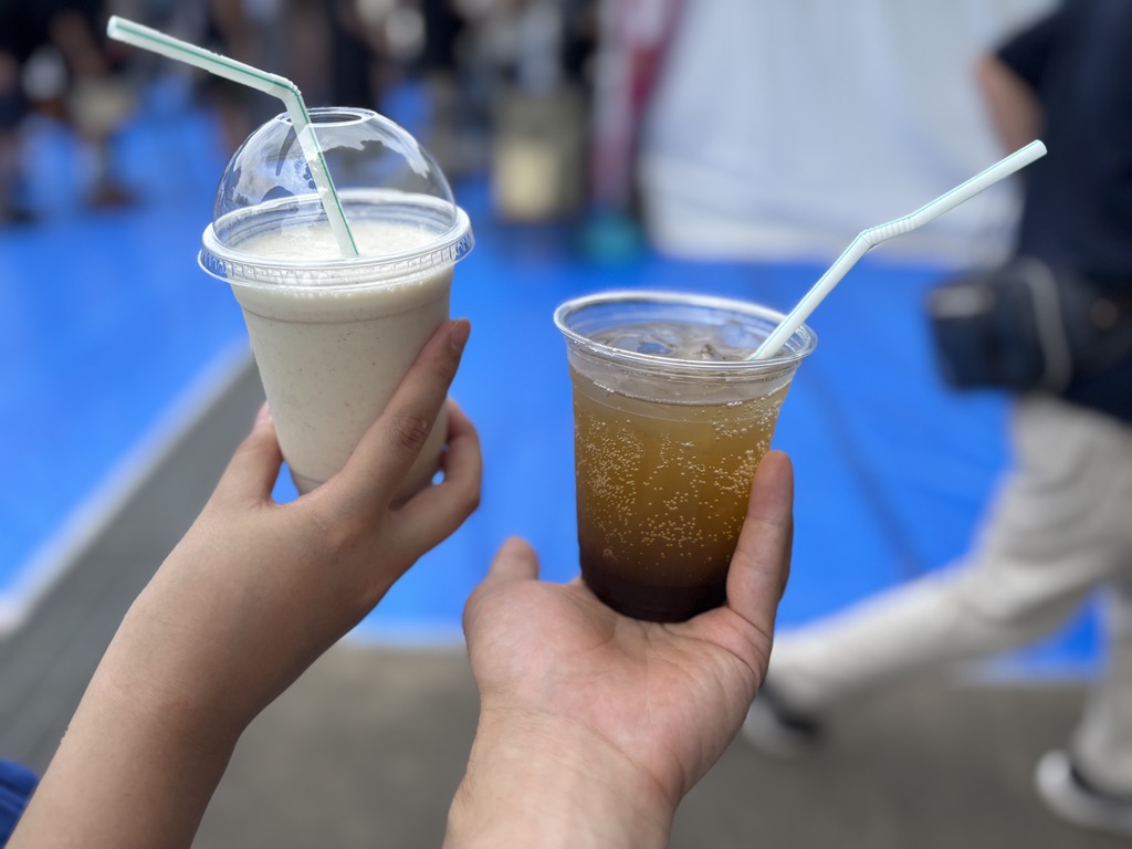 「左手にイチジクとくるみのミルクシェイク、右手にタマリンドレモネードソーダを持ち、会場を背景に撮影。」