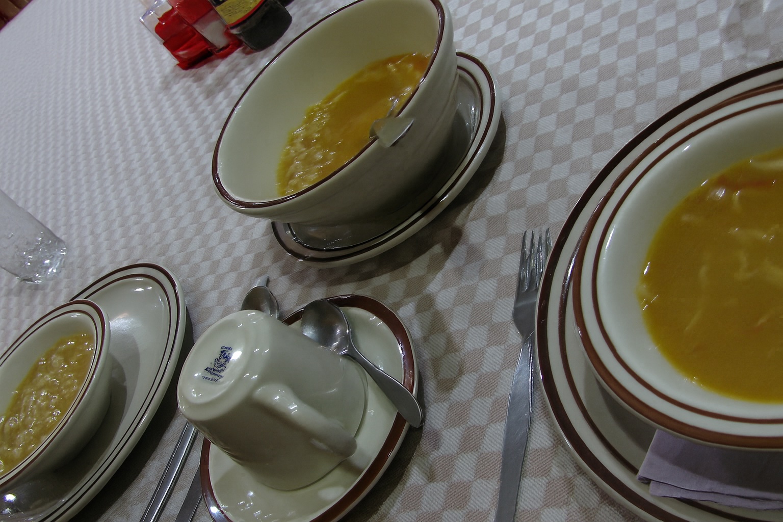 「茶色の縁取りがある器に盛られた黄金色のチキンヤクニ。白いテーブルクロスの上に並び、隣にはカップやスプーンが整えられている」