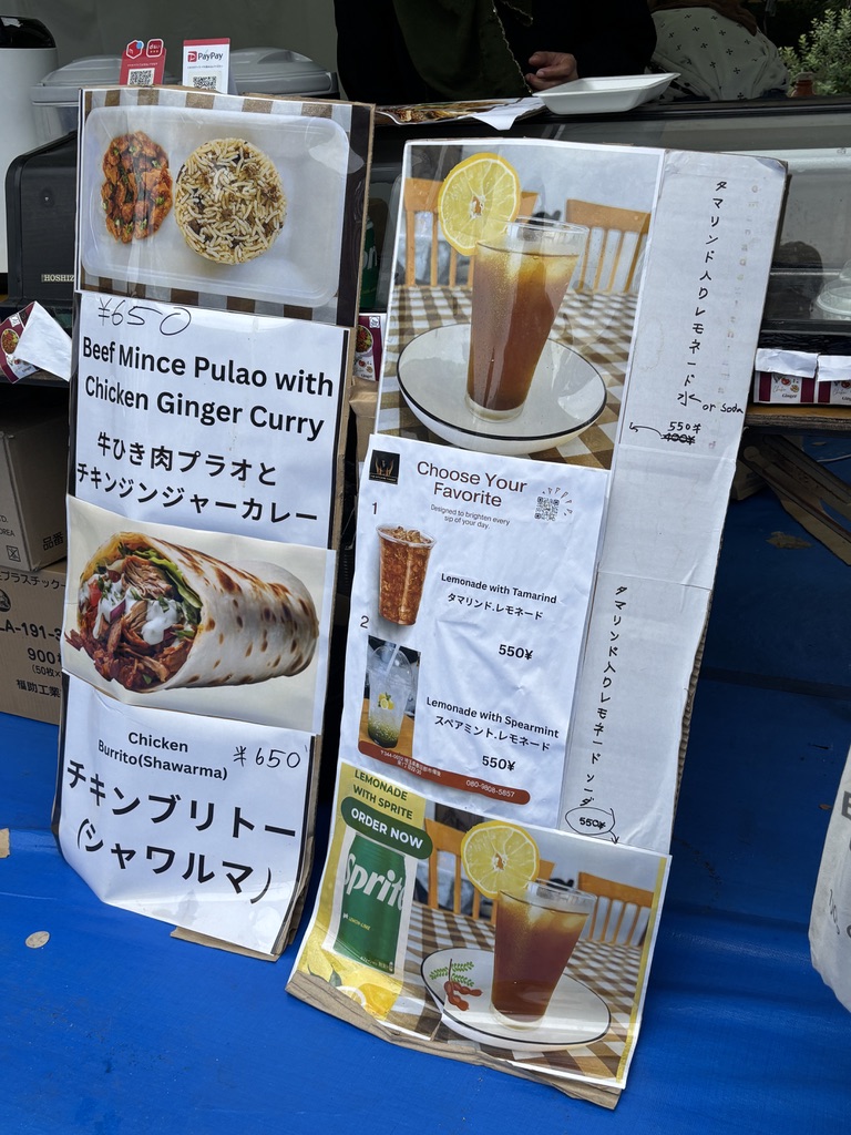 「上野公園のイベント会場に出店しているTwinflame Foodsの屋台。ビーフミンチプラオやチキンジンジャーカレー、シャワルマ、タマリンドレモネードなど多国籍料理を販売。カラフルなフルーツスムージーの看板も並ぶ。」
