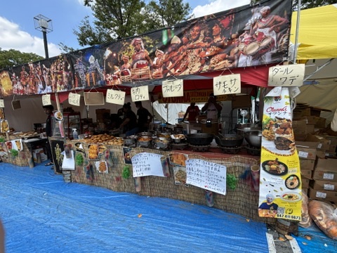 屋外イベントに出店しているスリランカ料理屋台。色鮮やかな民族舞踊の写真が屋台上部に飾られ、「ミルクティー」「牛肉ロティ」「フィッシュカレー」などの手書きメニュー札が並ぶ。前面には揚げ物やパン、カレー鍋がずらりと並び、スタッフが調理や接客をしている。青いビニールシートの床、背後に木々とテントが見える。