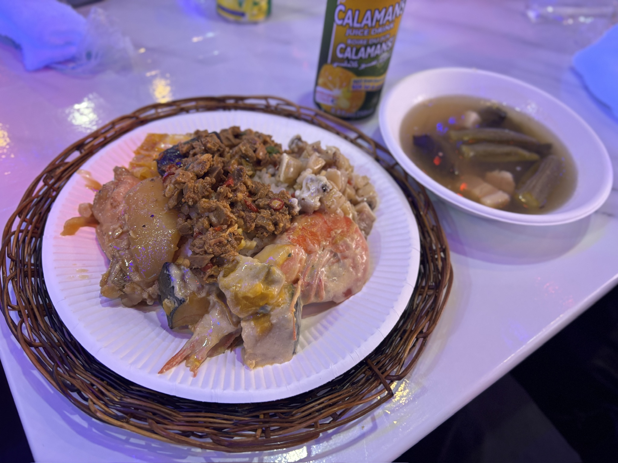 紙皿に盛られた色とりどりのフィリピン料理と、オクラ入りの酸味あるスープ。奥にはカラマンシージュース缶。