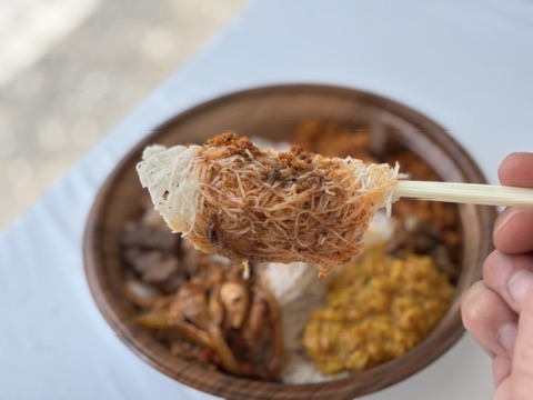 箸で持ち上げたインディアッパー（ストリングホッパー）に、スパイシーなマトンカレーを絡めた一口。奥に副菜が並ぶプレート。珍しい逸品。