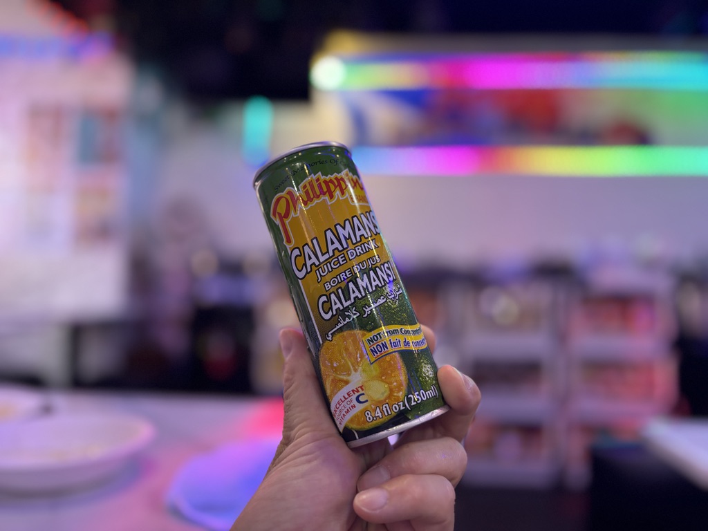 手に持ったフィリピンの柑橘系ジュース「カラマンシー」の缶。背景はネオンライトが彩る店内で、フィリピンらしいポップな雰囲気。