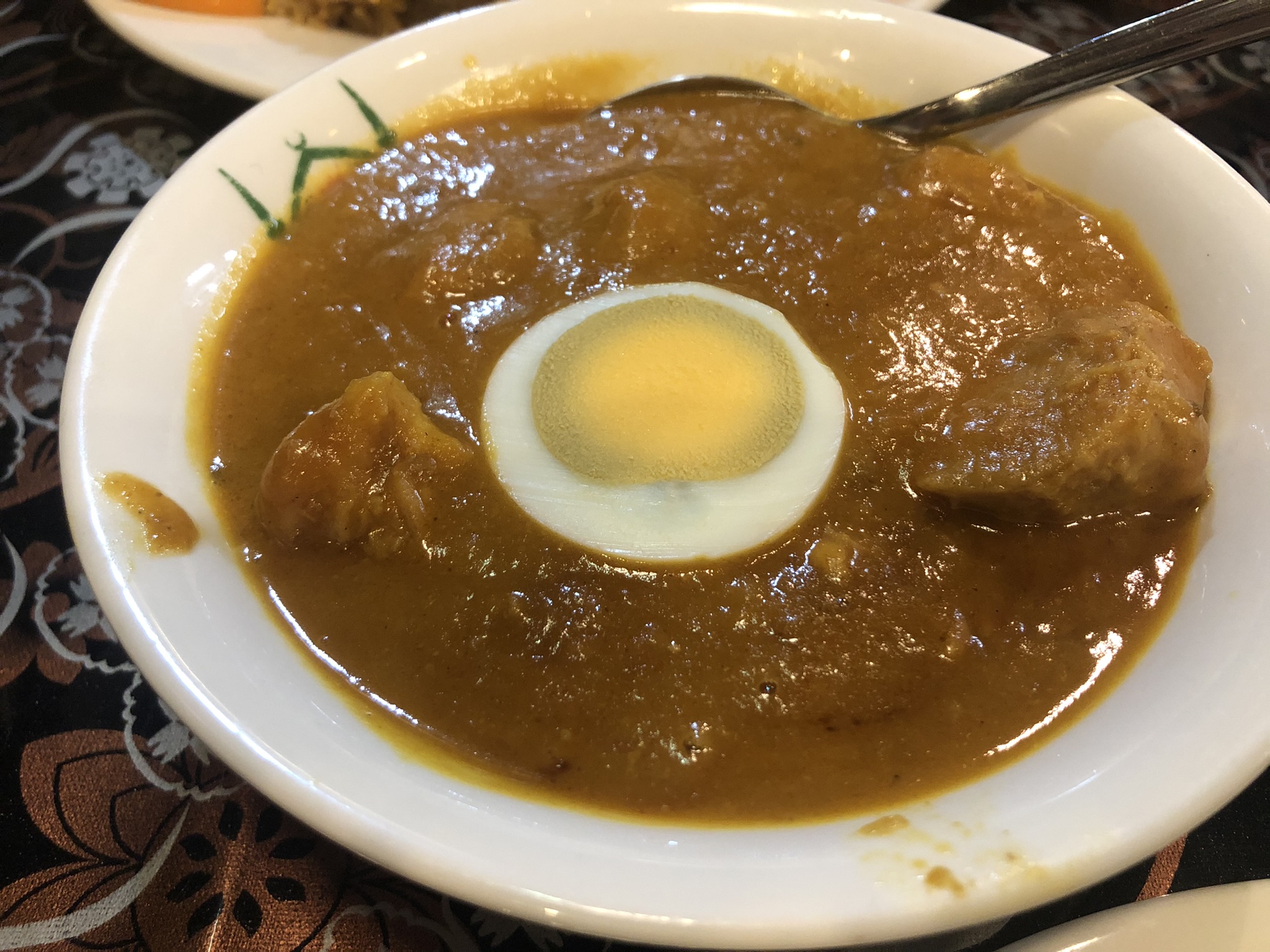 「白い深皿に盛られた濃厚なオレンジ色のチキンムグライ。中央には半分にカットされたゆで卵がのせられ、周囲には鶏肉の塊が見える。背景には褐色とベージュの花柄クロスが敷かれている」