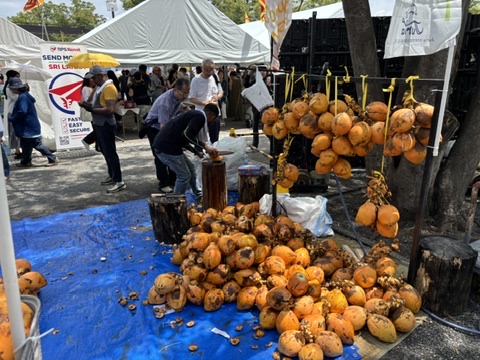 「スリランカフェスティバルの屋台で販売されるキングココナッツ。鮮やかなオレンジ色の実が束ねられ、店員がその場でカットしてジュースを提供している様子」